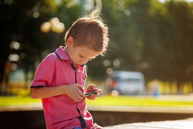 小さな子供は暖かい日当たりの良いダで遊ぶために彼の携帯電話に夢中