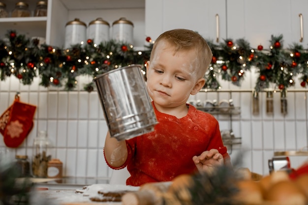 A little kid boy with iron sieve in his hand and dirty from flour helps mother to cook Christmas ginger biscuit in light kitchen