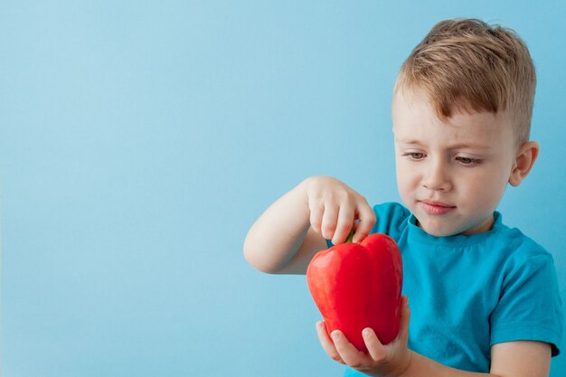 Little kid holding pepper in his hands on blue background. Vegan and healthy concept