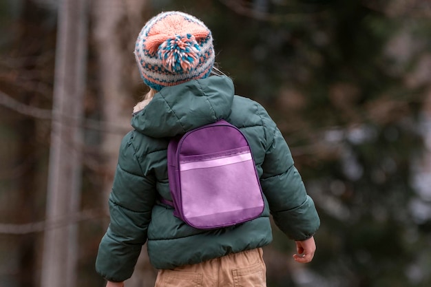 Photo little kid in knitted hat and jacket wears small backpack in pine forest child in fall in woods back view