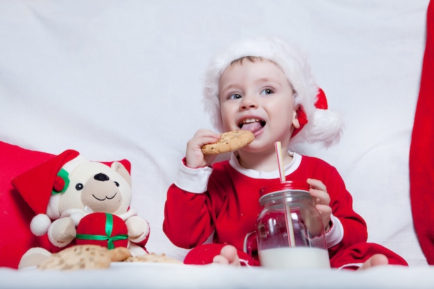 A little kid in a red cap eats a cookies and milk. Christmas photography of a baby in a red cap. New Year holidays and Christmas