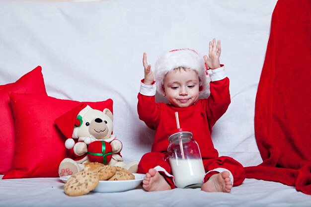 A little kid in a red cap eats a cookies and milk