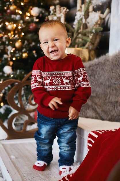 Little kid in red Christmas sweater posing on the stairs of the decorated for Christmas house.