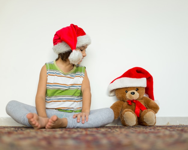 Little kid in Santa hat with cute teddy bear waiting at home for New year