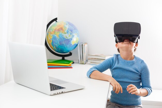 Little kid schoolgirl sit and study at white desk with pc laptop. School distance education at home during quarantine concept. Watching in headset, virtual glasses