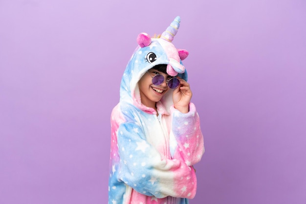 Little kid wearing a unicorn pajama isolated on purple background with glasses and happy