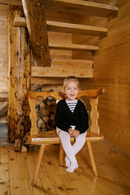 Little laughing girl sitting on a chair near the stairs in a wooden cottage