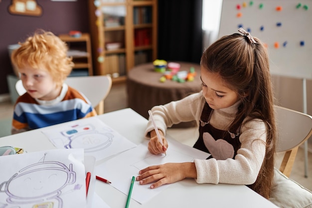 Little learner of nursery school drawing picture with crayons on paper while sitting by desk against