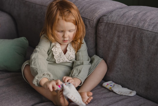 Little lightskinned cute girl with red hair in dress puts on socks while sitting on sofa at home Children independence concept