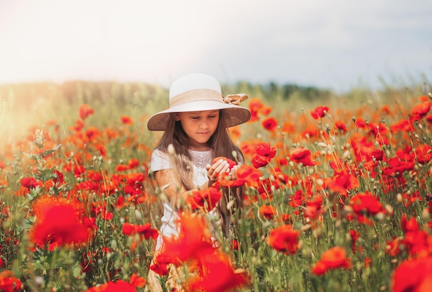 Little longhaired girl in hat posing at field of poppies with on summer sun