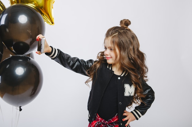 Little modern hipster girl in fashion clothes and baloons, Selfie. Take photo on telephone