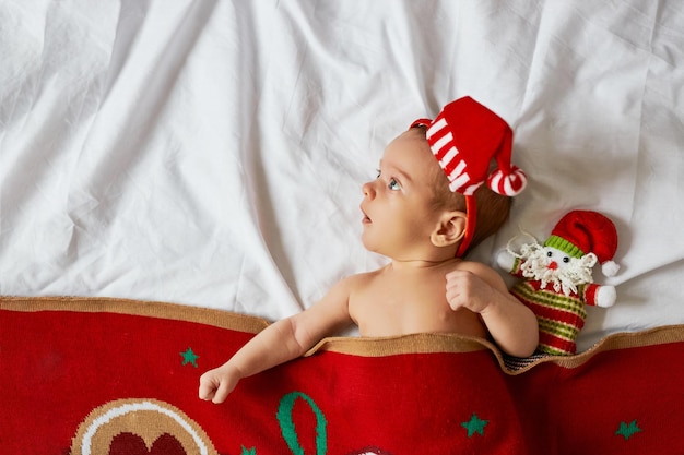 Little newborn baby, wearing Santa hat, lies under red Christmas blanket with toy. Top above view, x-mas home indoors