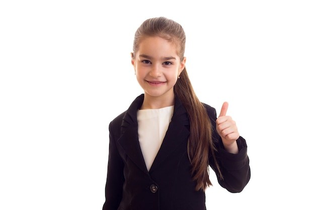 Little nice girl with long brown ponytail in white Tshirt and jacket showing thumb up in studio