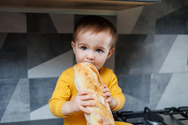 A little oneyearold boy is sitting in the kitchen and eating a long bread or baguette in the kitchen