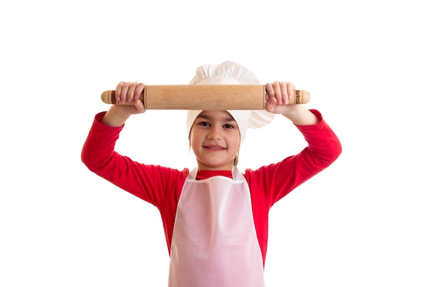 Little optimistic girl in red shirt with white apron and hat holding rolling pin in studio