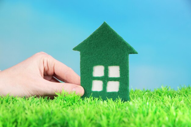 Little paper house in hand on green grass on blue sky background