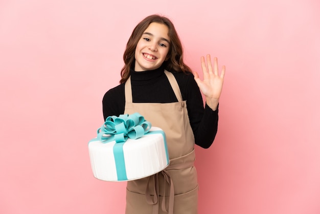 Little Pastry chef holding a big cake isolated on pink background saluting with hand with happy expression