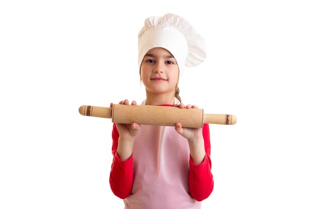 Little pleasant girl in red shirt with white apron and hat holding rolling pin and kitchen spatula