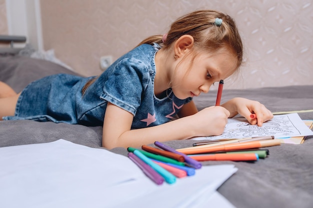 A little preschooler is lying on a soft bed and is engaged in coloring various pictures with felttip pens