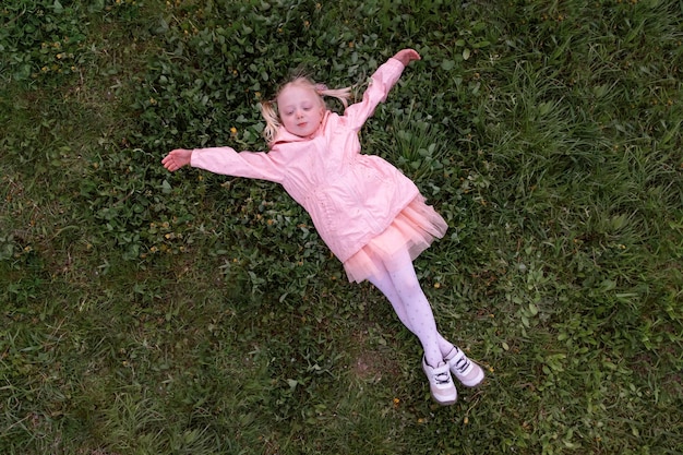 분홍색 드레스를 입은 작은 유치원 아이는 그녀의 팔을 펴고 휴식을 취하는 초록색 잔디에 누워 있습니다.