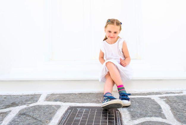 Little pretty blong girl in a white dress sitting outdoor on a street