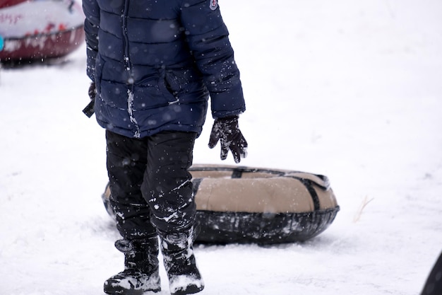 A little pretty boy in a dark overalls slides down an ice slide, winter children's games, a child in the park in winter. The boy climbs the slide with tubing