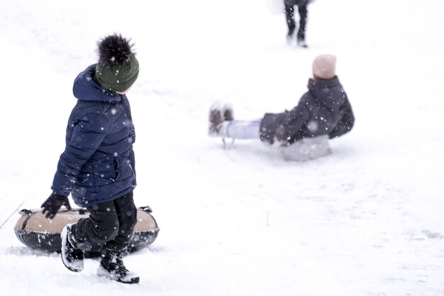 Симпатичный мальчик в темном комбинезоне скатывается по ледяной горке, зимние детские игры, зимой ребенок в парке. Мальчик поднимается по горке с трубкой