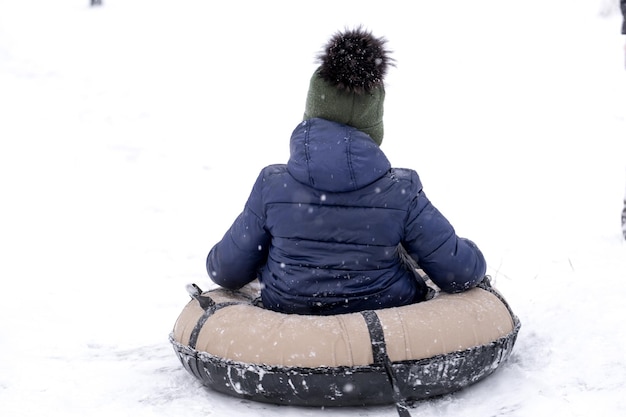 A little pretty boy in a dark overalls slides down an ice slide, winter children's games, a child in the park in winter. Little boy having extreme winter fun on tubing