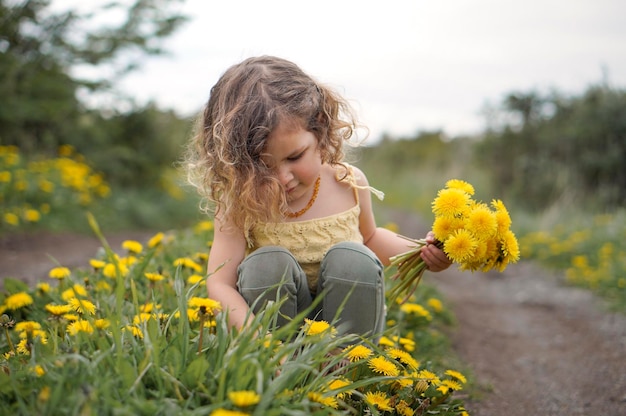 春にタンポポの黄色い花の花束を集める小さなかわいい巻き毛の女の子