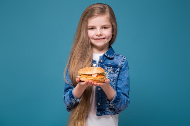 La piccola ragazza graziosa in giacca di jeans con lunghi capelli castani tiene un hamburger