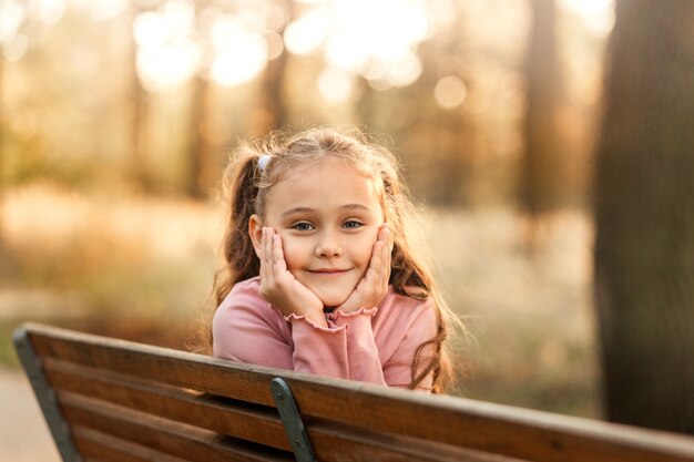 小さなかわいい女の子が秋の公園のベンチに座っています