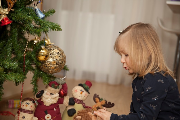 部屋のクリスマスツリーのそばに座っているクリスマスプレゼントを持った小さなかわいい女の子、新年のコンセプト