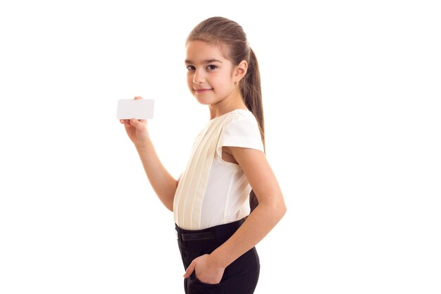 Маленькая красивая девочка с длинным коричневым хвостиком в белой футболке и черной юбке с белой карточкой
