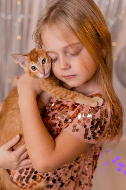 A little princess girl in an elegant dress holds a red kitten in her hands. The symbol of the year