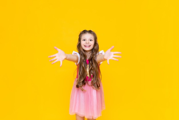 A little princess in a pink dress with curly hair smiles broadly and holds out her hands palms forward A beautiful charming child on a yellow isolated background