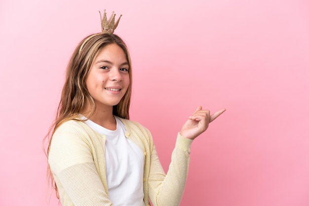 Little princess with crown isolated on pink background pointing finger to the side