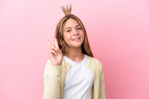 Little princess with crown isolated on pink background smiling and showing victory sign