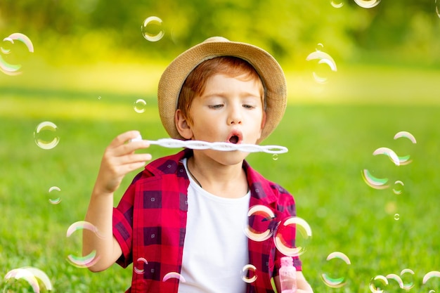 A little redhaired boy inflates soap bubbles on a green lawn in a hat and a red shirt in summer having fun playing