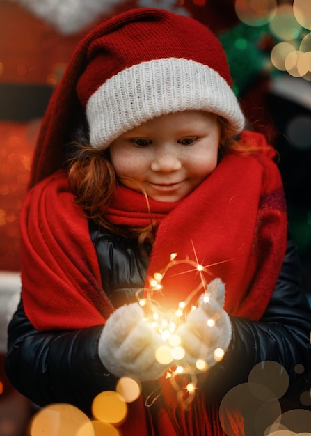 彼女の手にクリスマスライトを持っているサンタクロースの帽子をかぶった小さな赤毛の少女がクリスマスを待っています