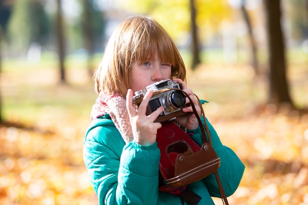 Foto piccola ragazza dai capelli rossi con una telecamera retro nel parco autunnale fotografo infantile