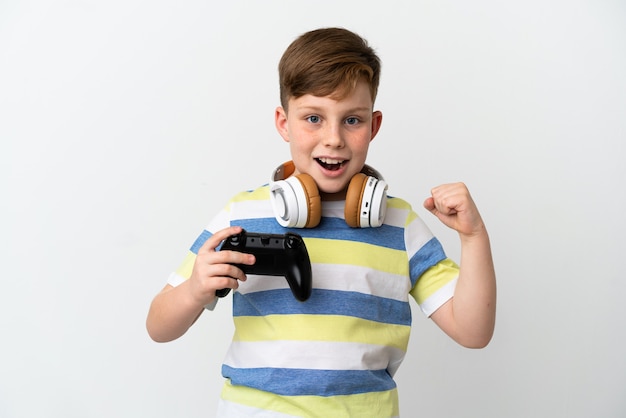 Little redhead boy holding a game pad isolated on white background celebrating a victory in winner position