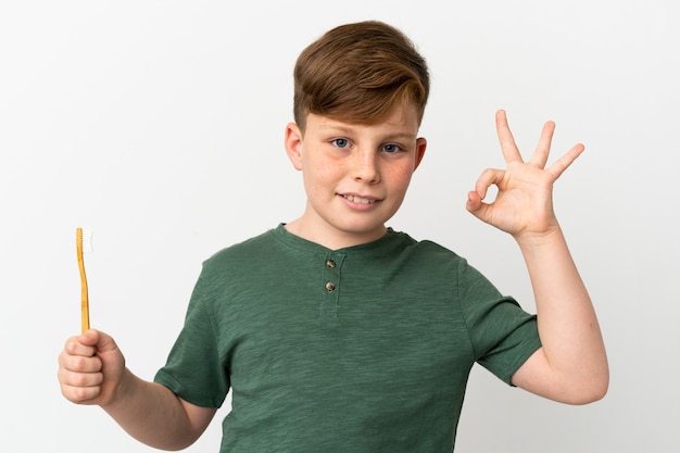 Little redhead boy holding a toothbrush isolated on white background showing ok sign with fingers