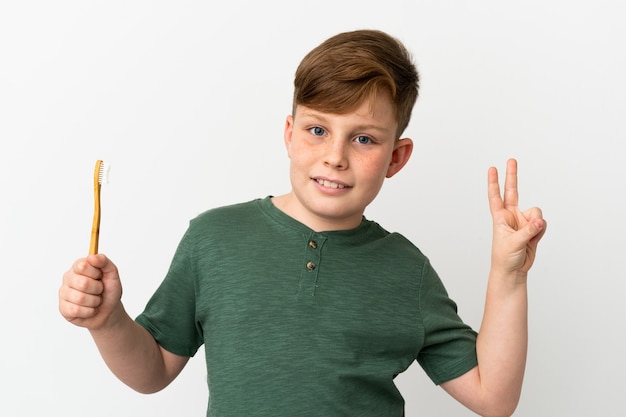 Little redhead boy holding a toothbrush isolated on white background smiling and showing victory sign