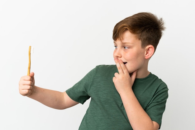 Little redhead boy holding a toothbrush isolated on white background with surprise and shocked facial expression