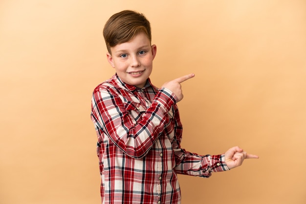 Little redhead boy isolated on beige background surprised and pointing side