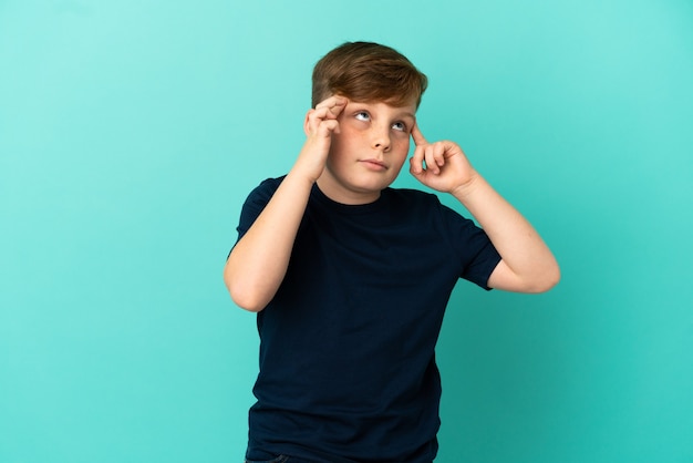 Little redhead boy isolated on blue background having doubts and thinking