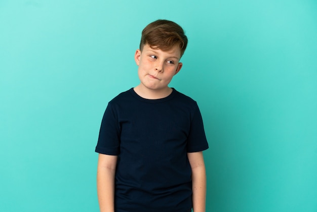 Little redhead boy isolated on blue background having doubts while looking up