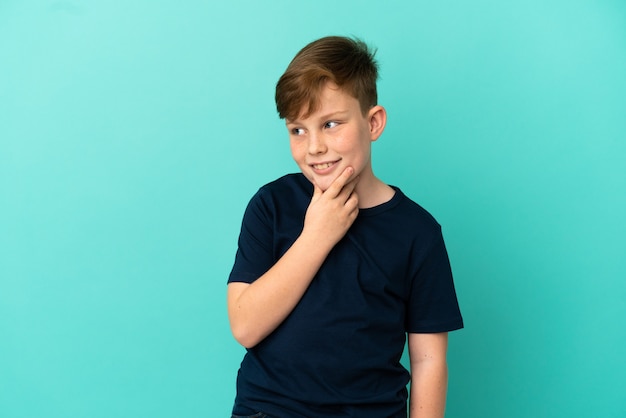 Ragazzino dai capelli rossi isolato su sfondo blu che guarda di lato e sorride