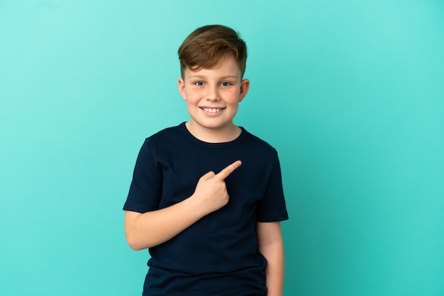 Little redhead boy isolated on blue background pointing to the side to present a product