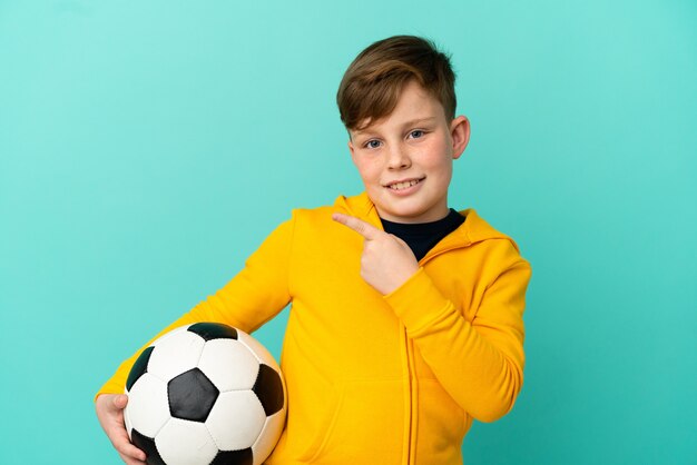 Little redhead boy isolated on blue background with soccer ball and pointing to the lateral